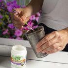 A person transplanting a small plant into a clear pot, with a container of takeroot rooting hormone nearby on a windowsill with flowers.