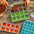 Person filling a seed starting tray with soil, with empty trays and plant markers on a wooden table.
